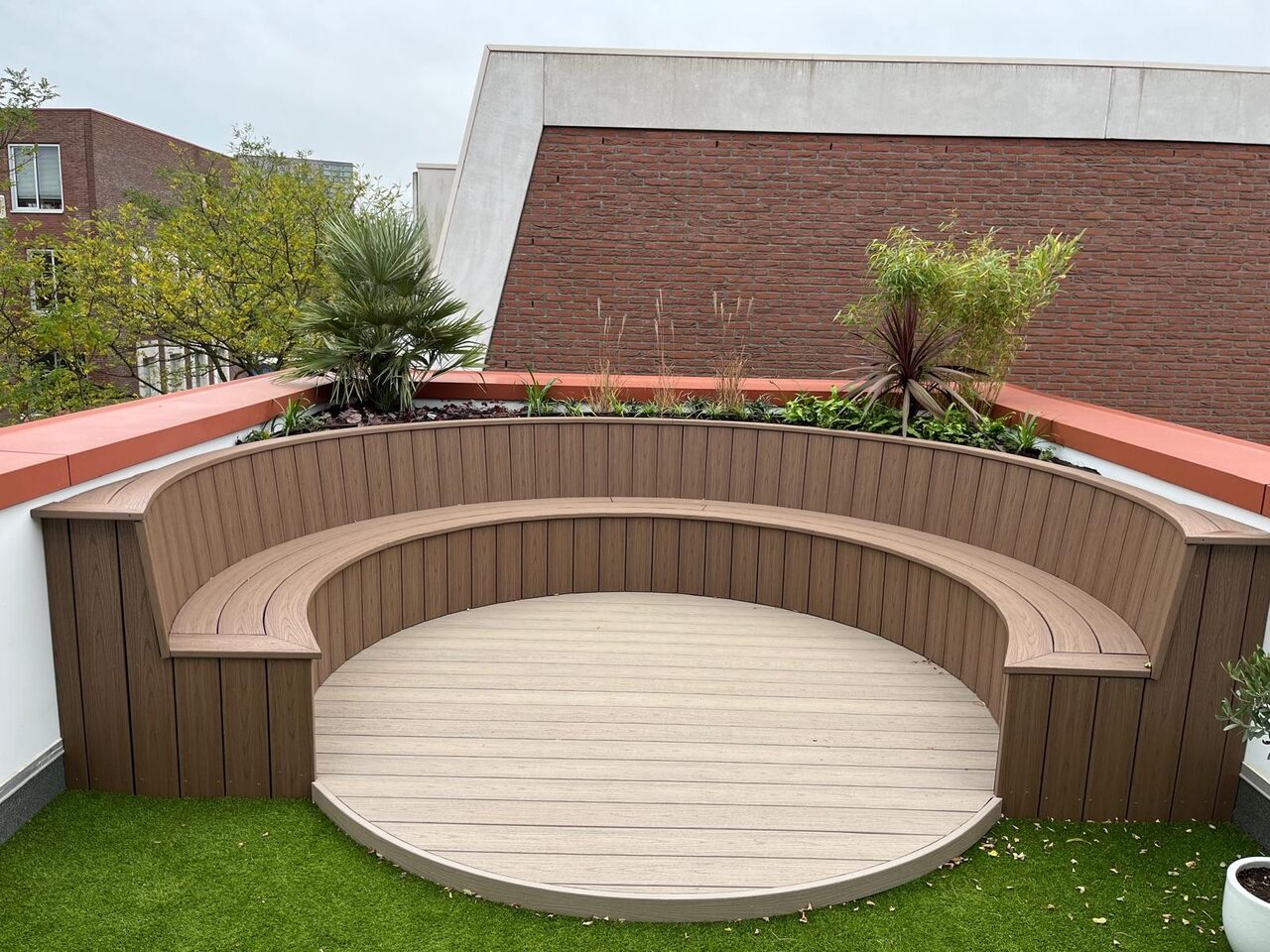 Dakterras met een ronde zitkuil van composiet vlonderdelen, gebogen bank, kunstgras en een border met palm en siergrassen