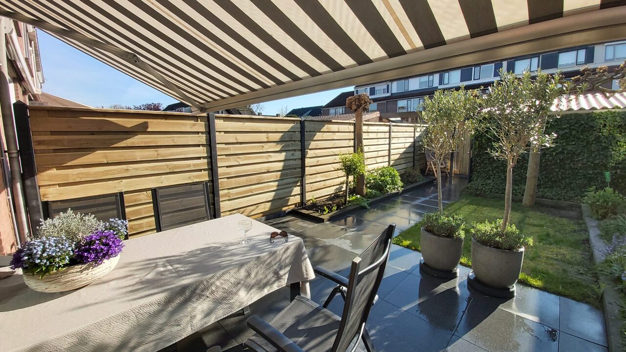 Terras onder een gestreept uitvalscherm met gedekte eettafel en olijfbomen in betonnen potten